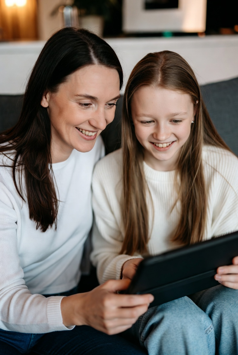 Mutter sitzt neben der Tochter, beide schauen auf ein Tablet und spielen Strategy Mission.